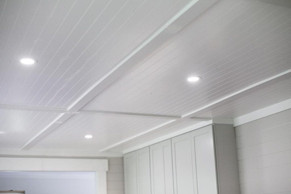White beadboard ceiling with recessed lights and structural beams in a bright room.
