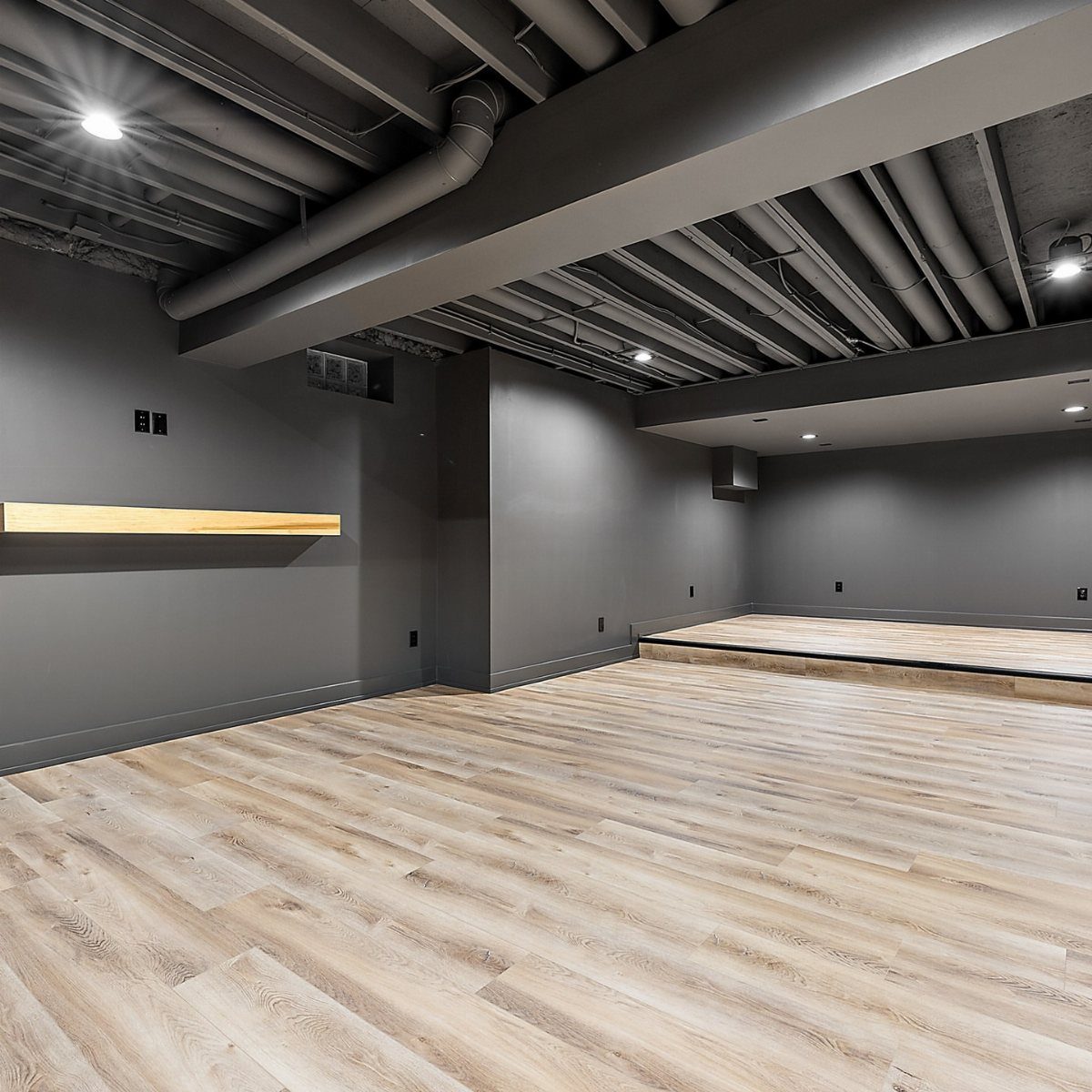 Exposed basement ceiling painted black with industrial pipes and light wood flooring.