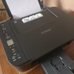 Black Canon PIXMA printer and scanner on a wooden desk with white paper.