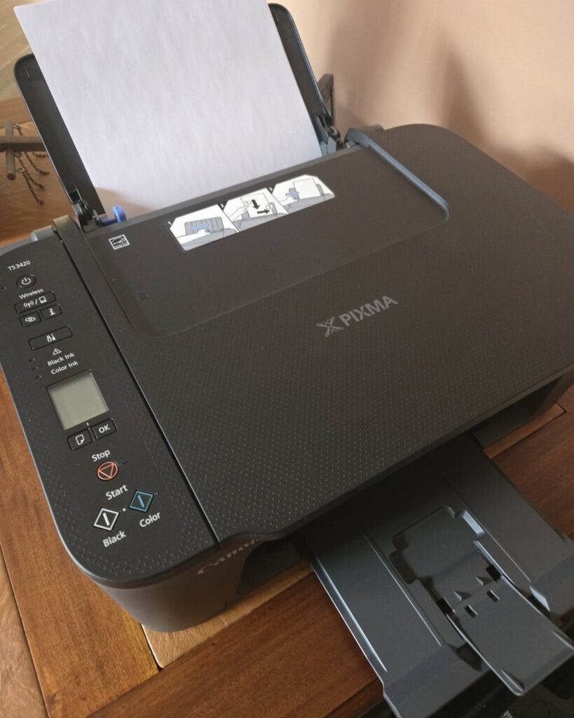 Black Canon PIXMA printer and scanner on a wooden desk with white paper.