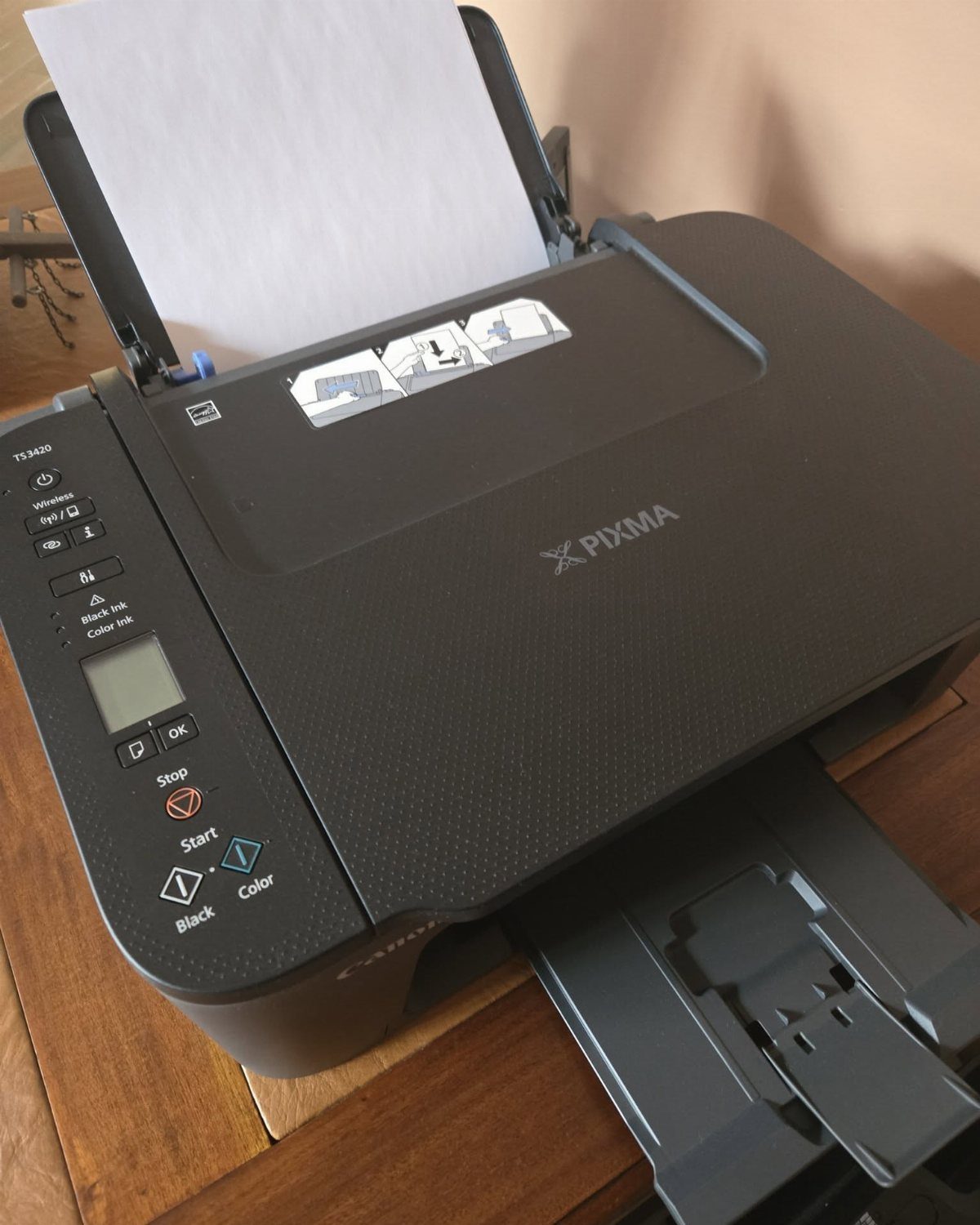 Black Canon PIXMA printer and scanner on a wooden desk with white paper.