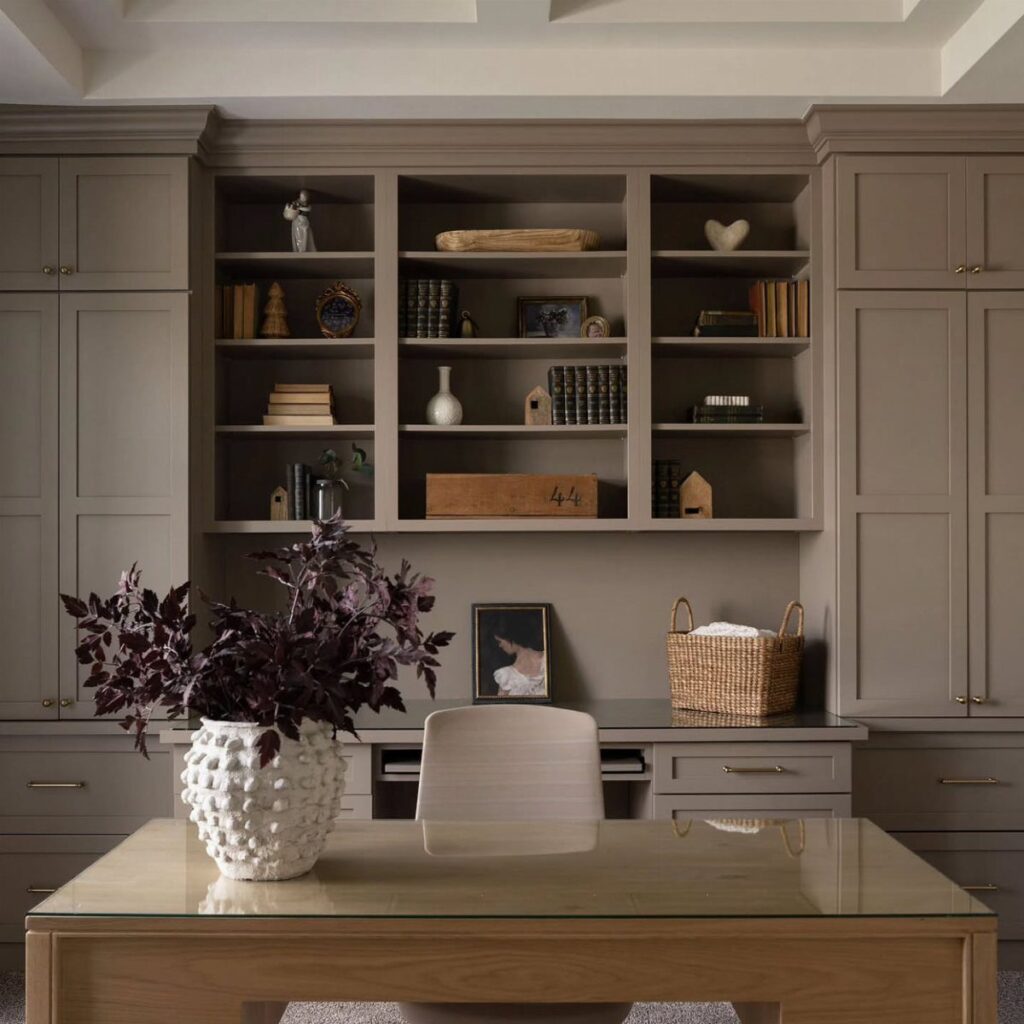 Gray built-in office cabinetry and shelving with a wooden glass-top table and purple foliage.