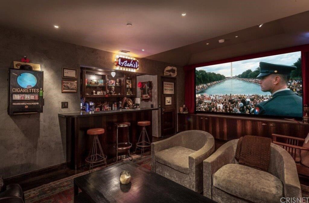 Man cave with a dark wood bar, grey armchairs, and a large movie screen.