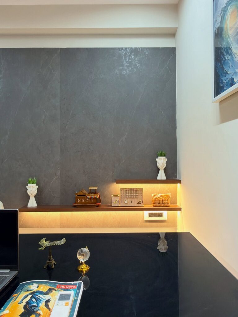 Home office with a dark grey wall, lit floating shelf, and black desk.