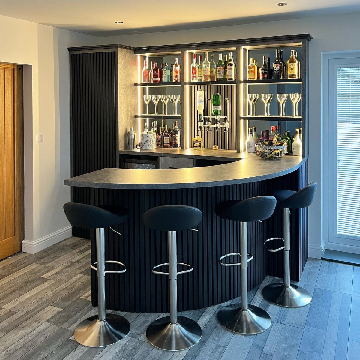 Curved black home bar with illuminated shelves, liquor bottles, and four modern bar stools.