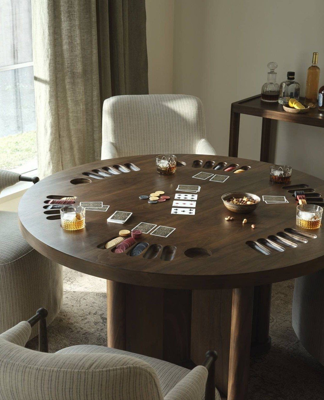Round dark wood poker table with cards, chips, and drinks in a bright room
