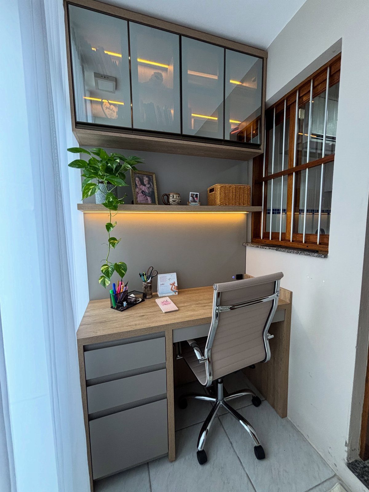 Compact home office with light wood desk, ergonomic chair, and overhead glass cabinets.